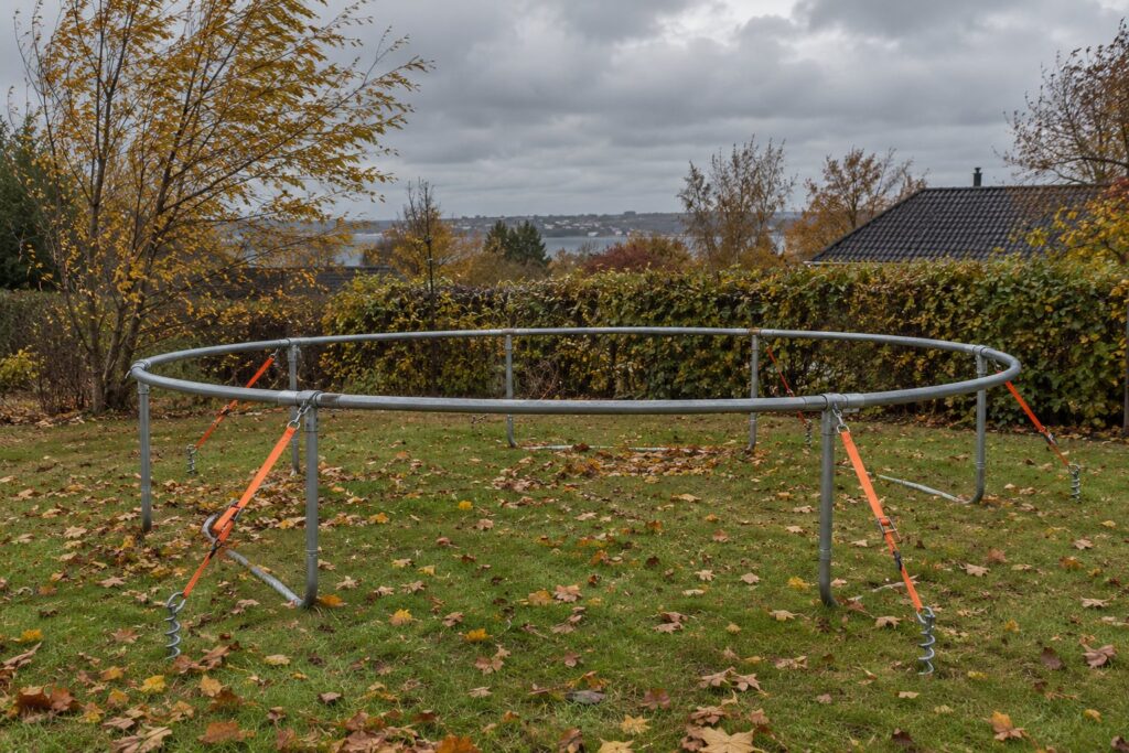 Trampolinramme uden dug og net spændt fast til græsplænen med stropper og skruepløkker i en dansk have en blæsende efterårsdag